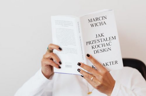 Anonymous female in white shirt with gold chain on neck reading interesting book covering face while sitting on black chair